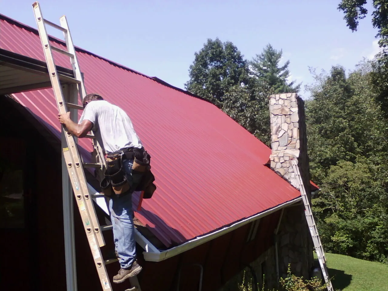 Roofer on a ladder beside a red metal roof and stone chimney — Soffit & Fascia Repair by Roofing Brothers in Lake Havasu City
